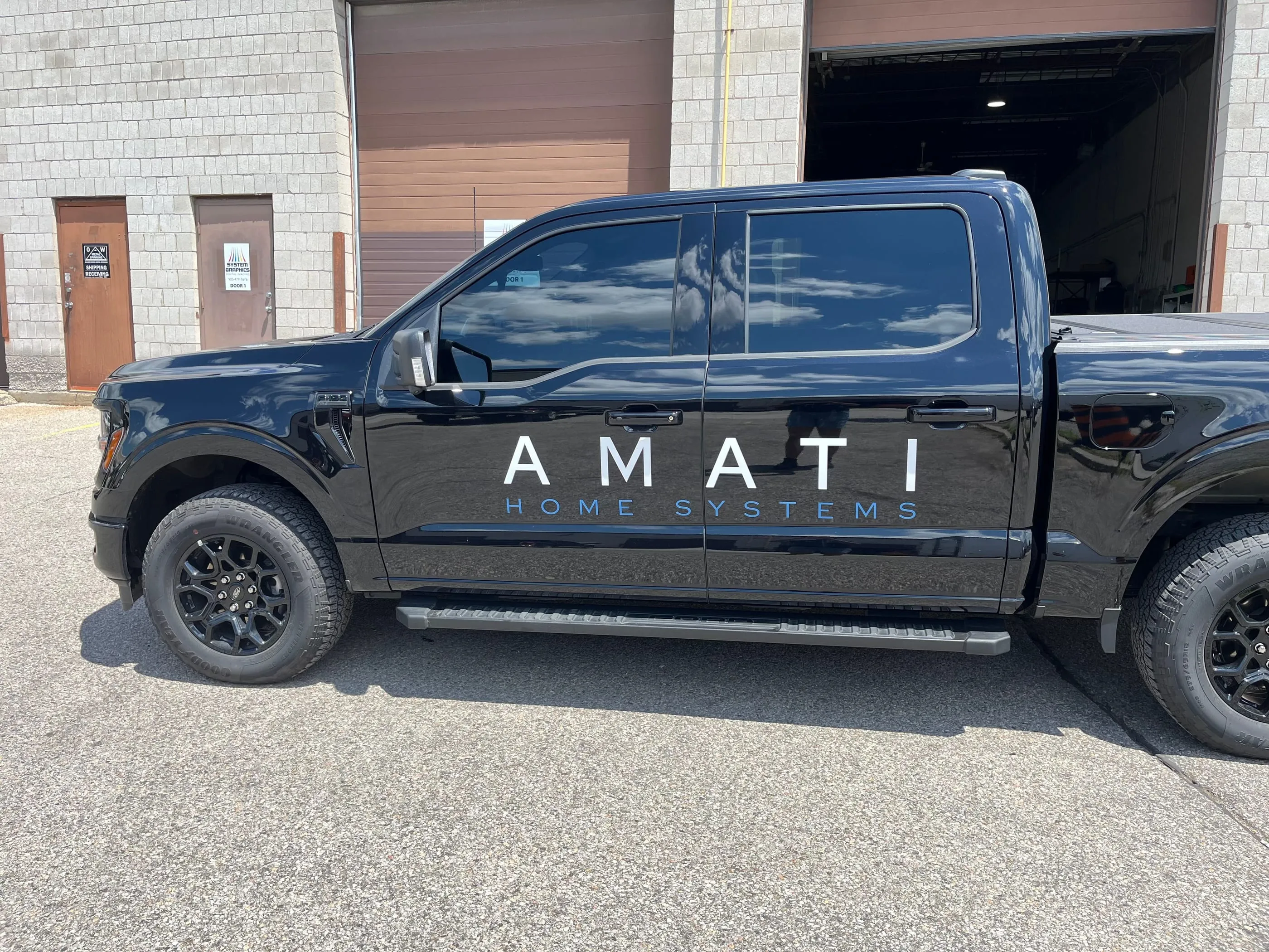 Black pickup truck with white Amati Home Systems door lettering, parked outside a shop