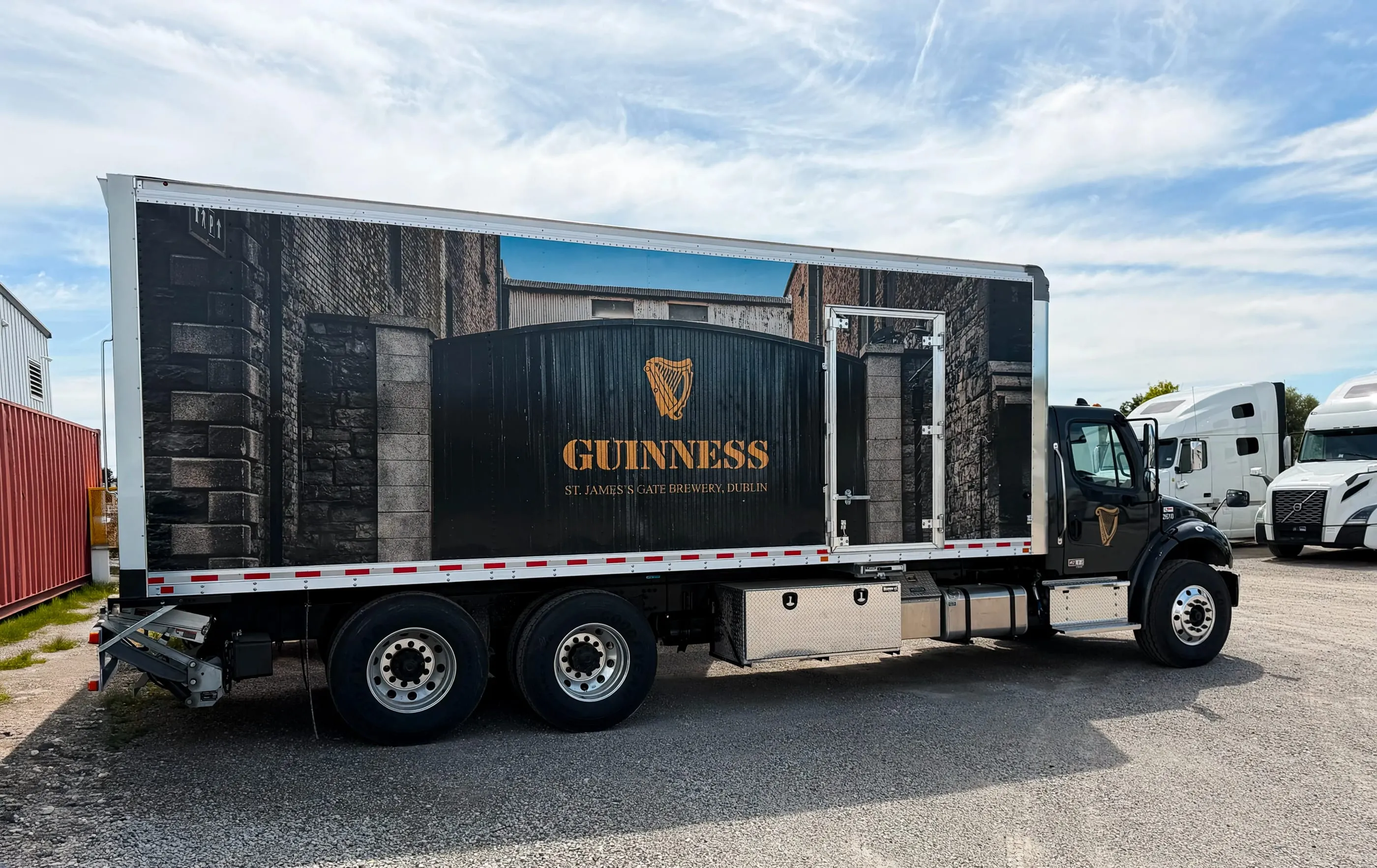 Guinness box truck wrapped with a stone brewery gate scene, parked in a yard under a blue sky
