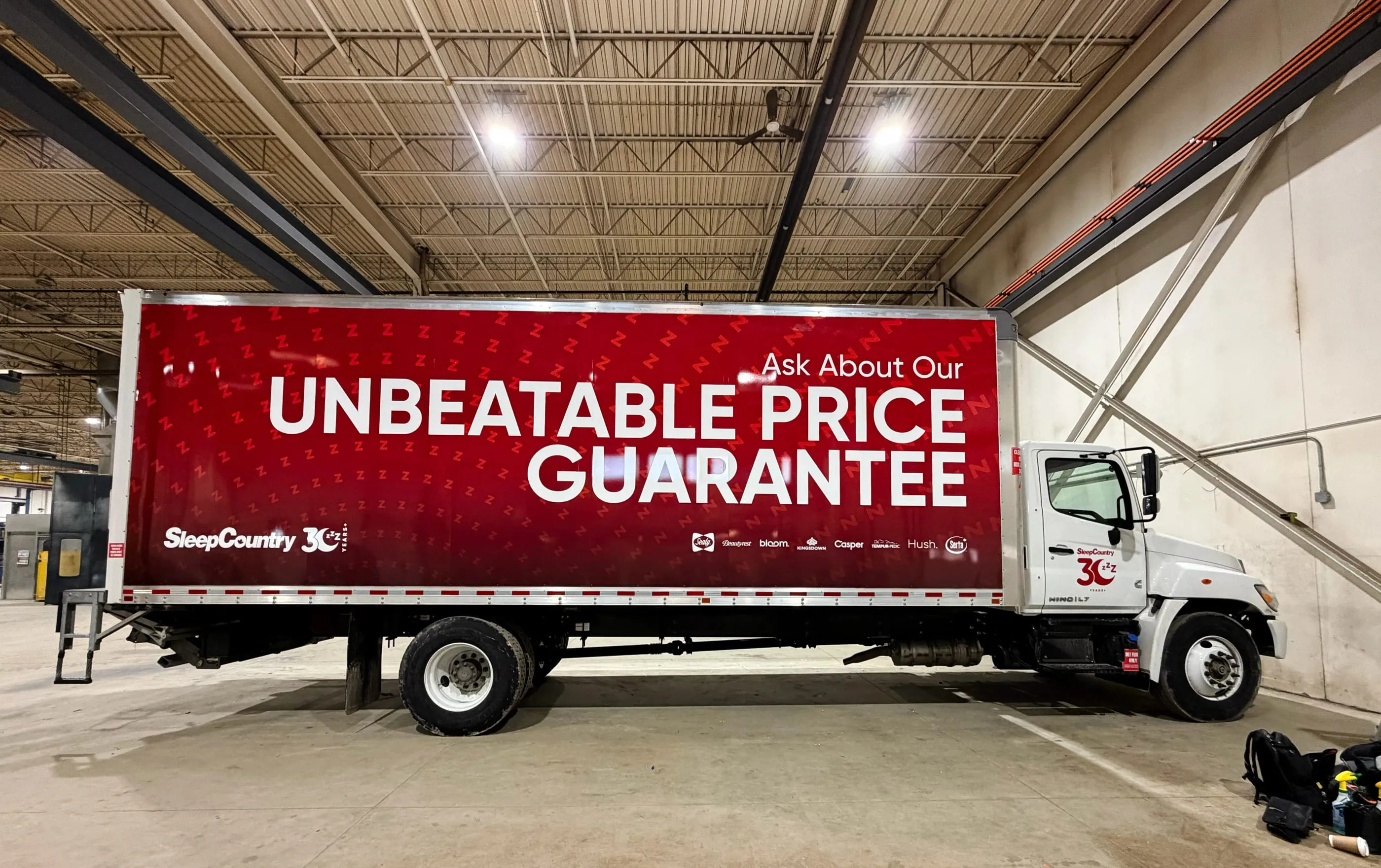 Sleep Country box truck with red full-side graphics reading “Unbeatable Price Guarantee”, in the shop bay